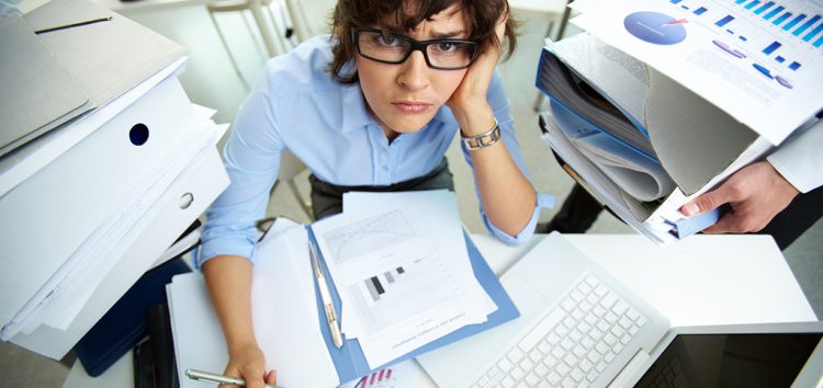 Woman working too hard in an office with files and paperwork all over her desk.