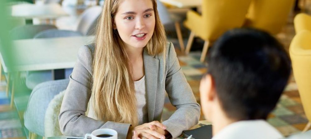 Portrait of two modern young women discussing work sitting at table in cafe during business meeting or job interview