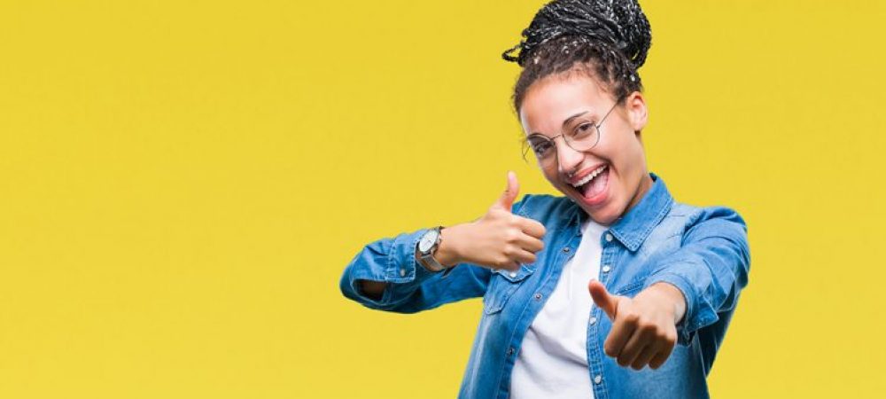 Young braided hair african american girl wearing glasses over isolated background approving doing positive gesture with hand, thumbs up smiling and happy for success. Looking at the camera, winner gesture.